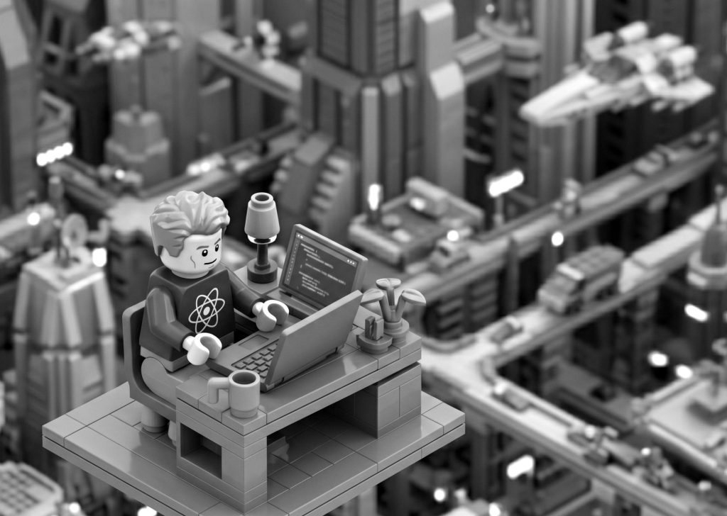 Black and white Lego developer working on a laptop above a futuristic city skyline, wearing an atom symbol shirt representing advanced react native mobile application development services.
