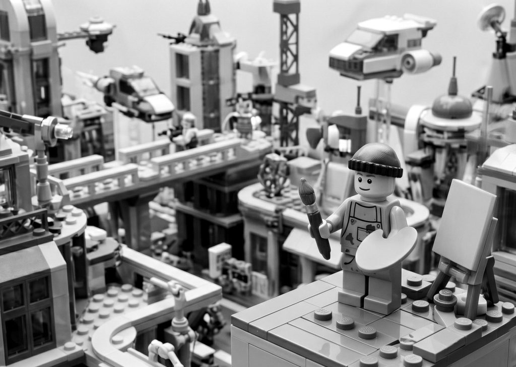 Black and white Lego artist holding a paintbrush and palette next to a blank canvas overlooking a futuristic Lego city, symbolizing the creative process from scratch and answering what is custom software development.