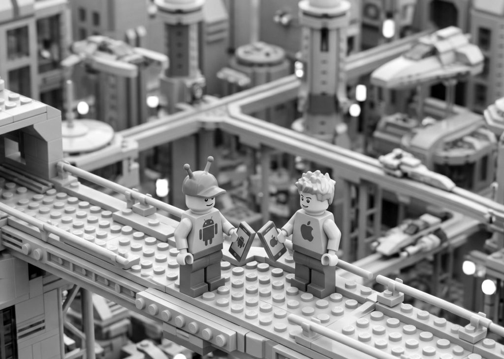 Black and white Lego figures representing Android and iOS platforms bumping smartphones on a futuristic city bridge, symbolizing the comparison of native vs. cross-platform app development.