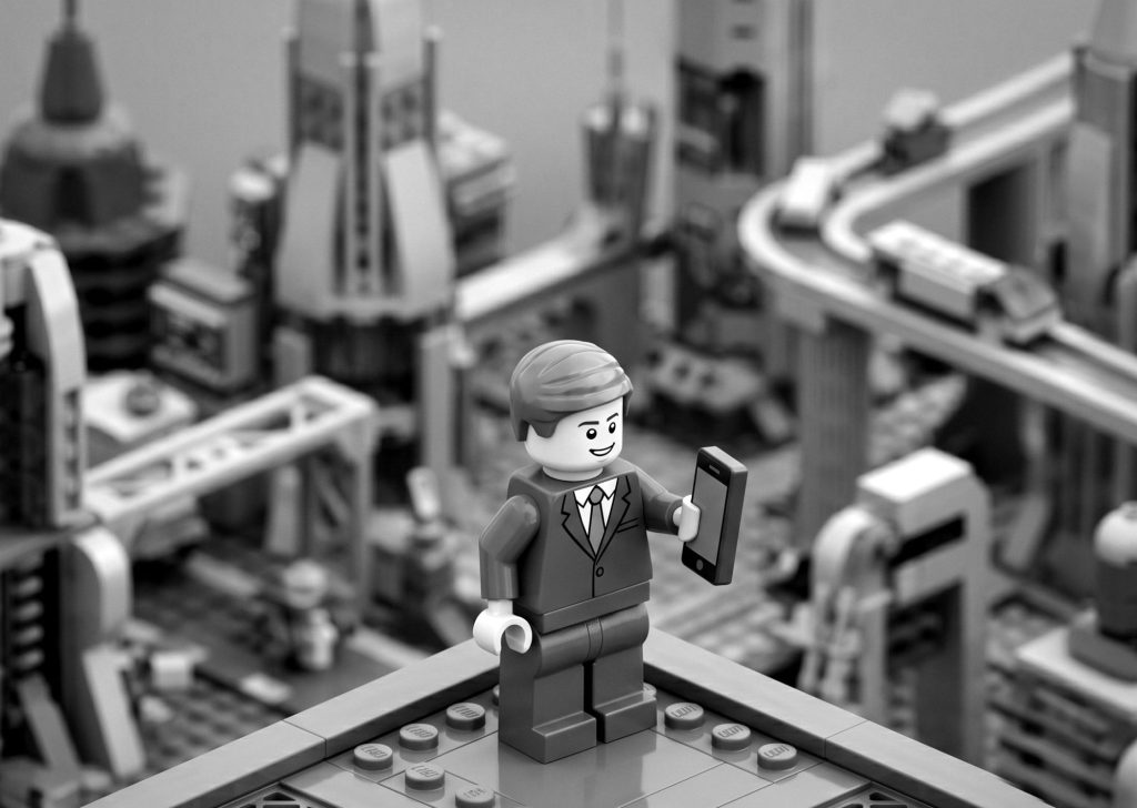 Black and white Lego businessman holding a smartphone overlooking a modern Lego city skyline, representing mobile connectivity and cross-platform app development.